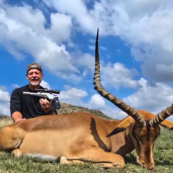 Hunter kneeling behind a long-horned impala ram with a scoped hunting handgun