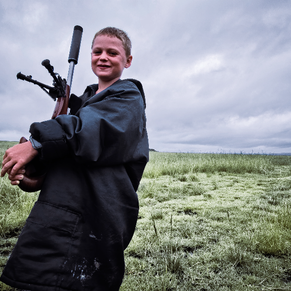 Boy in an oversized oilskin coat carrying a suppressed rifle across wet grass